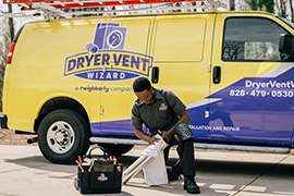 Dryer Vent Wizard professional kneels while cleaning dryer vent duct in front of company van.