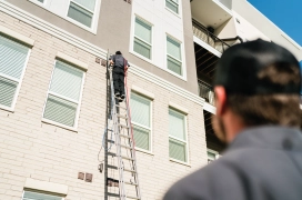 Dryer vent  cleaning professional on ladder outside of apartment complex and cleaning dryer ducts while his coworker watches from the ground.