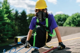 Dryer vent cleaning professional wearing hard hat and mask sits on roof of apartment complex and cleans dryer vent ducts.